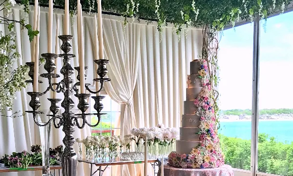 A tall, multi-tiered wedding cake decorated with cascading pink and white flowers, displayed on a table next to a large silver candelabra and floral arrangements.