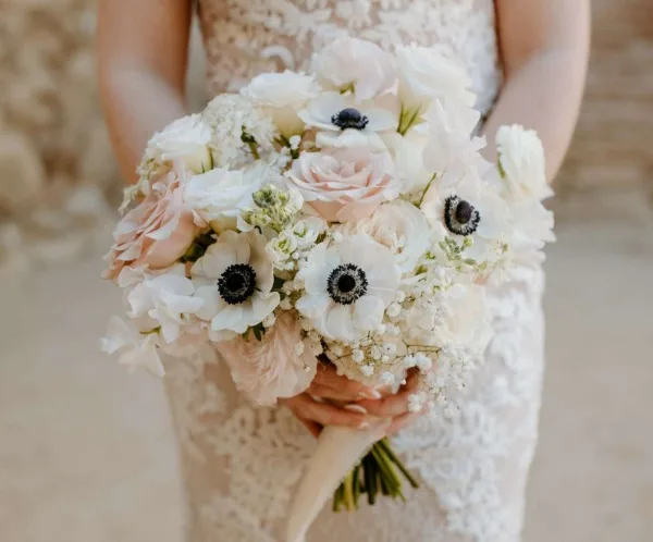 A bride in a lace dress holding a large, custom wedding bouquet featuring white and blush roses, anemones, and baby's breath.