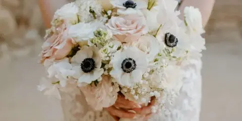 A bride in a lace dress holding a large, custom wedding bouquet featuring white and blush roses, anemones, and baby's breath.