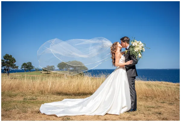 A bride and groom kissing on a grassy cliff overlooking the ocean with the bride's veil blowing in the wind.