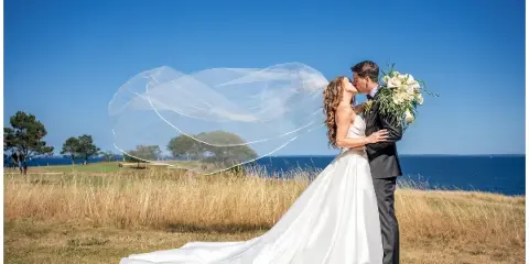A bride and groom kissing on a grassy cliff overlooking the ocean with the bride's veil blowing in the wind.