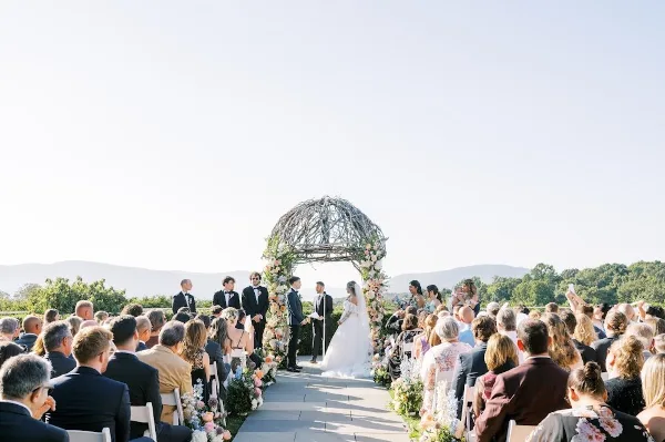 A wedding ceremony in progress with a bride and groom at a floral-decorated arch, surrounded by seated guests and floral aisle arrangements.
