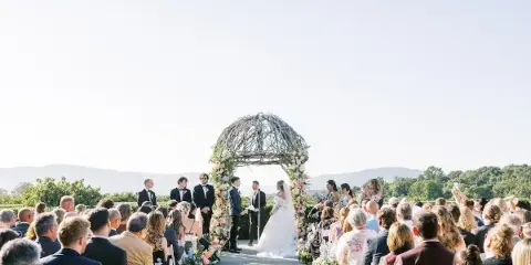 A wedding ceremony in progress with a bride and groom at a floral-decorated arch, surrounded by seated guests and floral aisle arrangements.