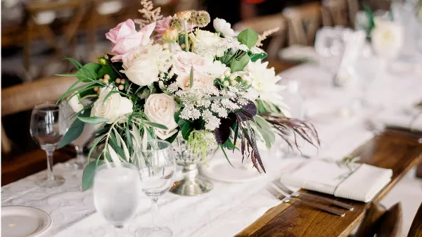 A close-up of a wedding table setting featuring a lush floral arrangement with pink and white roses, glassware, and white linens on a wooden table.