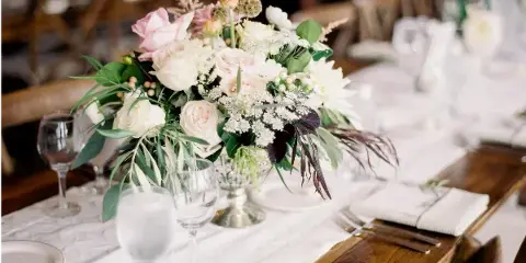 A close-up of a wedding table setting featuring a lush floral arrangement with pink and white roses, glassware, and white linens on a wooden table.
