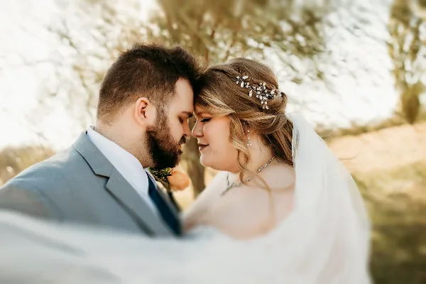 A bride and groom touching foreheads in an outdoor setting, wearing wedding attire.
