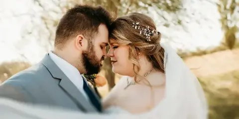A bride and groom touching foreheads in an outdoor setting, wearing wedding attire.