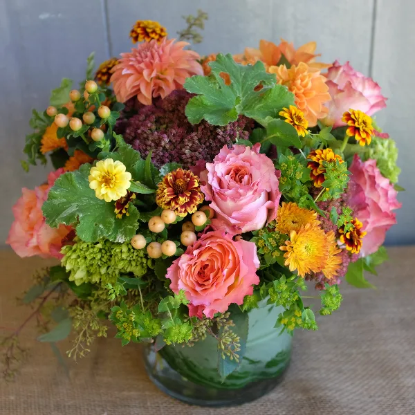 A vibrant floral arrangement in a glass vase featuring pink and orange roses, dahlias, and greenery.