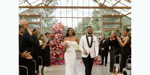 A bride and groom walking down the aisle in a glass-walled venue with a large floral backdrop and guests clapping.
