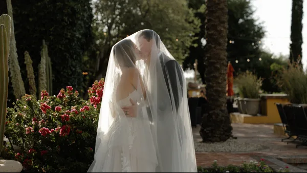 A bride and groom kissing under a veil in an outdoor garden setting.