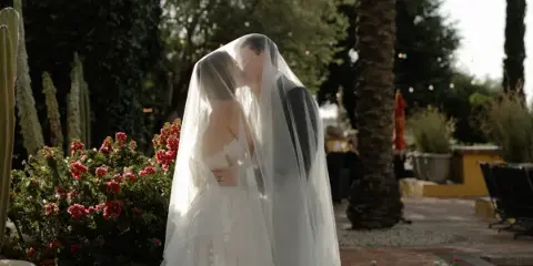 A bride and groom kissing under a veil in an outdoor garden setting.