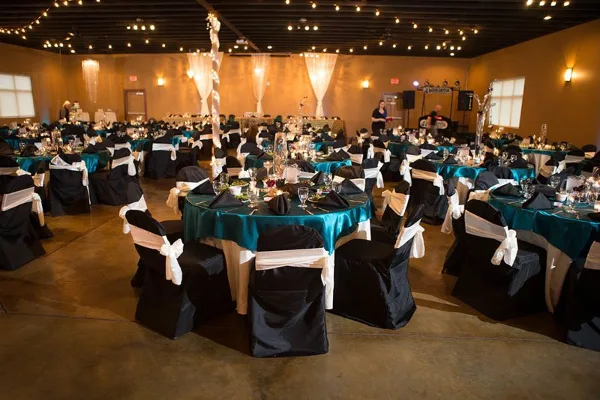 Banquet event venue with round tables in teal tablecloths, black chair covers with white sashes, string lights on the ceiling, and a band setup in the background.