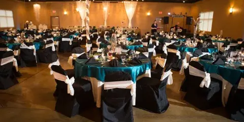 Banquet event venue with round tables in teal tablecloths, black chair covers with white sashes, string lights on the ceiling, and a band setup in the background.