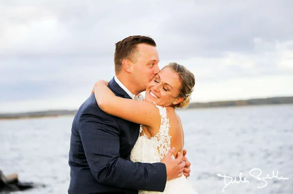 A bride and groom embracing and smiling outdoors near a body of water, with a photographer's signature in the corner.