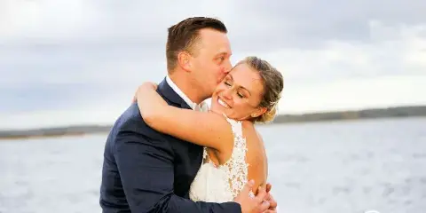 A bride and groom embracing and smiling outdoors near a body of water, with a photographer's signature in the corner.