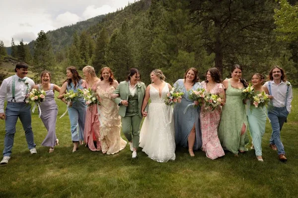A wedding party including the bride and bridesmaids posing outdoors in a grassy field with a forested mountain background.