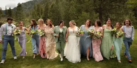 A wedding party including the bride and bridesmaids posing outdoors in a grassy field with a forested mountain background.
