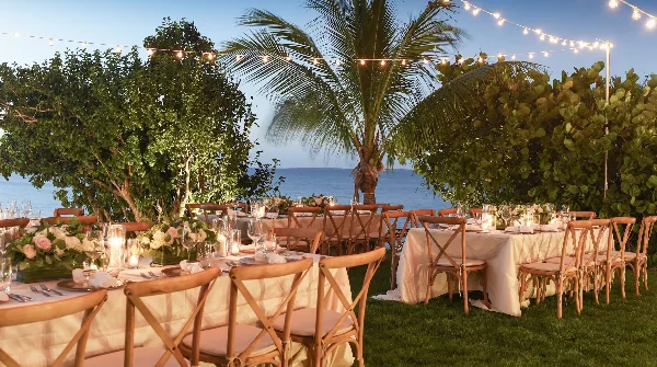 An outdoor wedding reception venue featuring long tables with white linens, floral arrangements, and string lights set against a tropical ocean view.