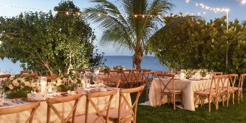 An outdoor wedding reception venue featuring long tables with white linens, floral arrangements, and string lights set against a tropical ocean view.