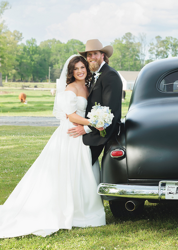 A bride and groom posing together outdoors next to a vintage black car.