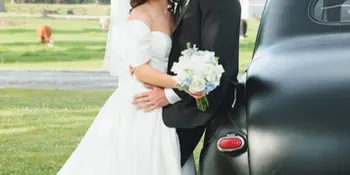 A bride and groom posing together outdoors next to a vintage black car.