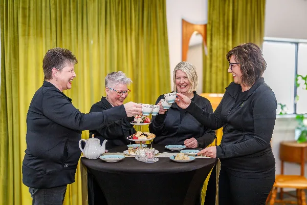 Four women in black attire toasting with teacups at a table featuring a tiered stand of tea sandwiches and scones.