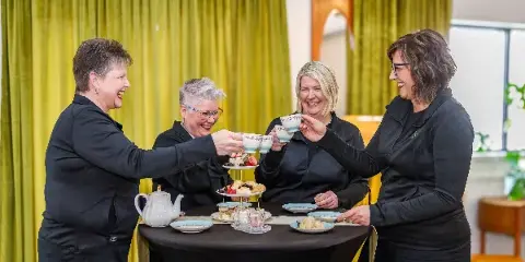 Four women in black attire toasting with teacups at a table featuring a tiered stand of tea sandwiches and scones.
