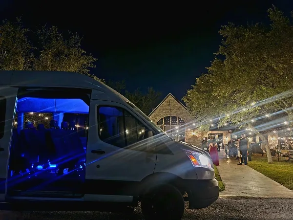 A shuttle van with blue interior lighting parked at a venue with string lights and guests at night.