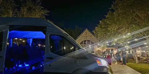 A shuttle van with blue interior lighting parked at a venue with string lights and guests at night.
