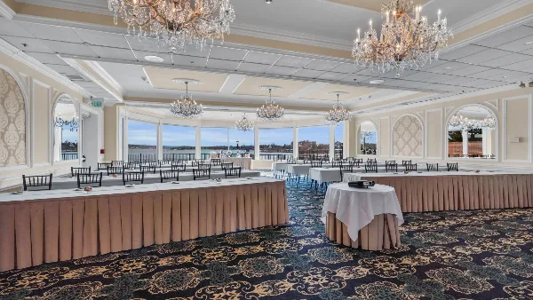 A spacious ballroom with chandeliers and tables set for an event, featuring large windows with a view of the water.