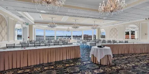 A spacious ballroom with chandeliers and tables set for an event, featuring large windows with a view of the water.