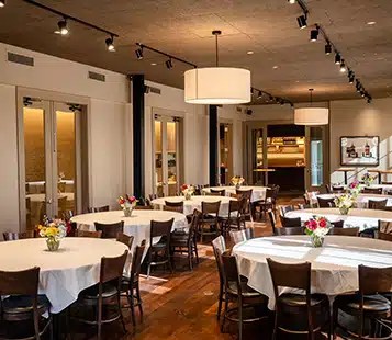 A private event venue with round tables set for a dinner, featuring white tablecloths and floral centerpieces.