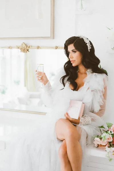 A bride with styled hair and makeup, wearing a white gown and headpiece, holding a champagne glass in a bridal suite setting.