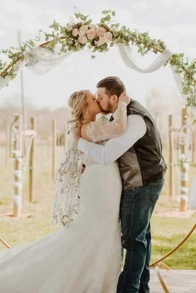 A bride and groom sharing a kiss under a floral arch during an outdoor wedding ceremony in a vineyard.