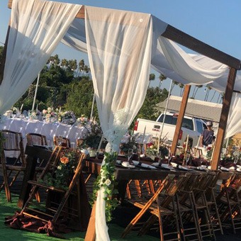 An outdoor wedding setup featuring a wooden canopy draped with white fabric, surrounded by tables and chairs.