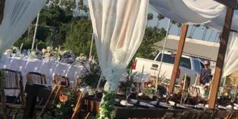 An outdoor wedding setup featuring a wooden canopy draped with white fabric, surrounded by tables and chairs.