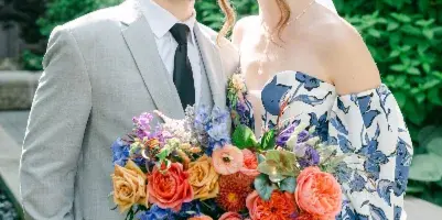 A bride and groom standing together outdoors, with the bride holding a large, colorful bouquet of flowers.