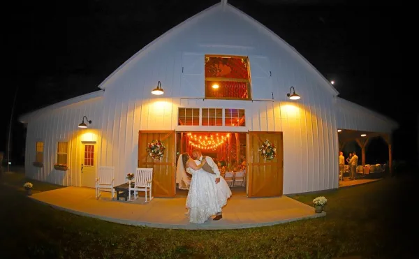 A white barn venue at night with a bride and groom standing on the porch in front of the open entrance.