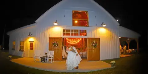 A white barn venue at night with a bride and groom standing on the porch in front of the open entrance.
