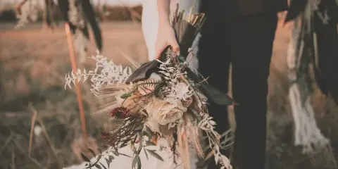 A bride and groom holding a rustic wedding bouquet in a field.