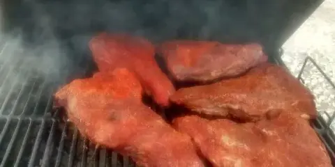 Large cuts of seasoned meat smoking on a grill grate with visible smoke.