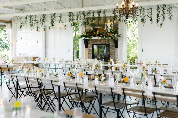 A wedding reception hall with long wooden tables, floral centerpieces, and hanging greenery.