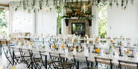 A wedding reception hall with long wooden tables, floral centerpieces, and hanging greenery.