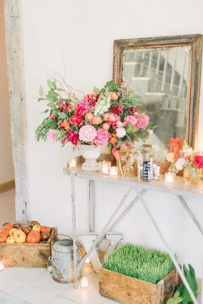 A rustic wedding floral arrangement with pink and orange blooms on a metal console table, accompanied by candles and a wooden crate of tomatoes.
