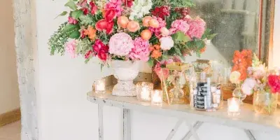 A rustic wedding floral arrangement with pink and orange blooms on a metal console table, accompanied by candles and a wooden crate of tomatoes.