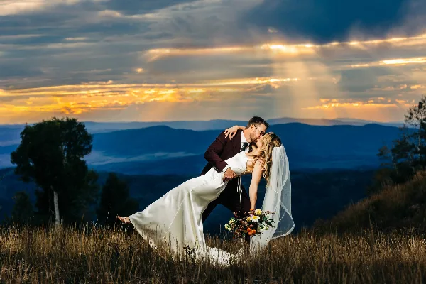 A bride and groom sharing a kiss on a grassy hilltop with a dramatic sunset and mountain range in the background.