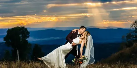A bride and groom sharing a kiss on a grassy hilltop with a dramatic sunset and mountain range in the background.