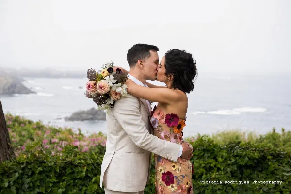 A bride and groom kissing outdoors with a wedding bouquet, set against a coastal background.