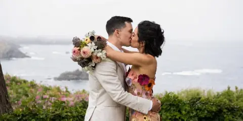 A bride and groom kissing outdoors with a wedding bouquet, set against a coastal background.