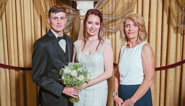 A bride and groom standing with a female wedding officiant in front of a gold curtain backdrop.
